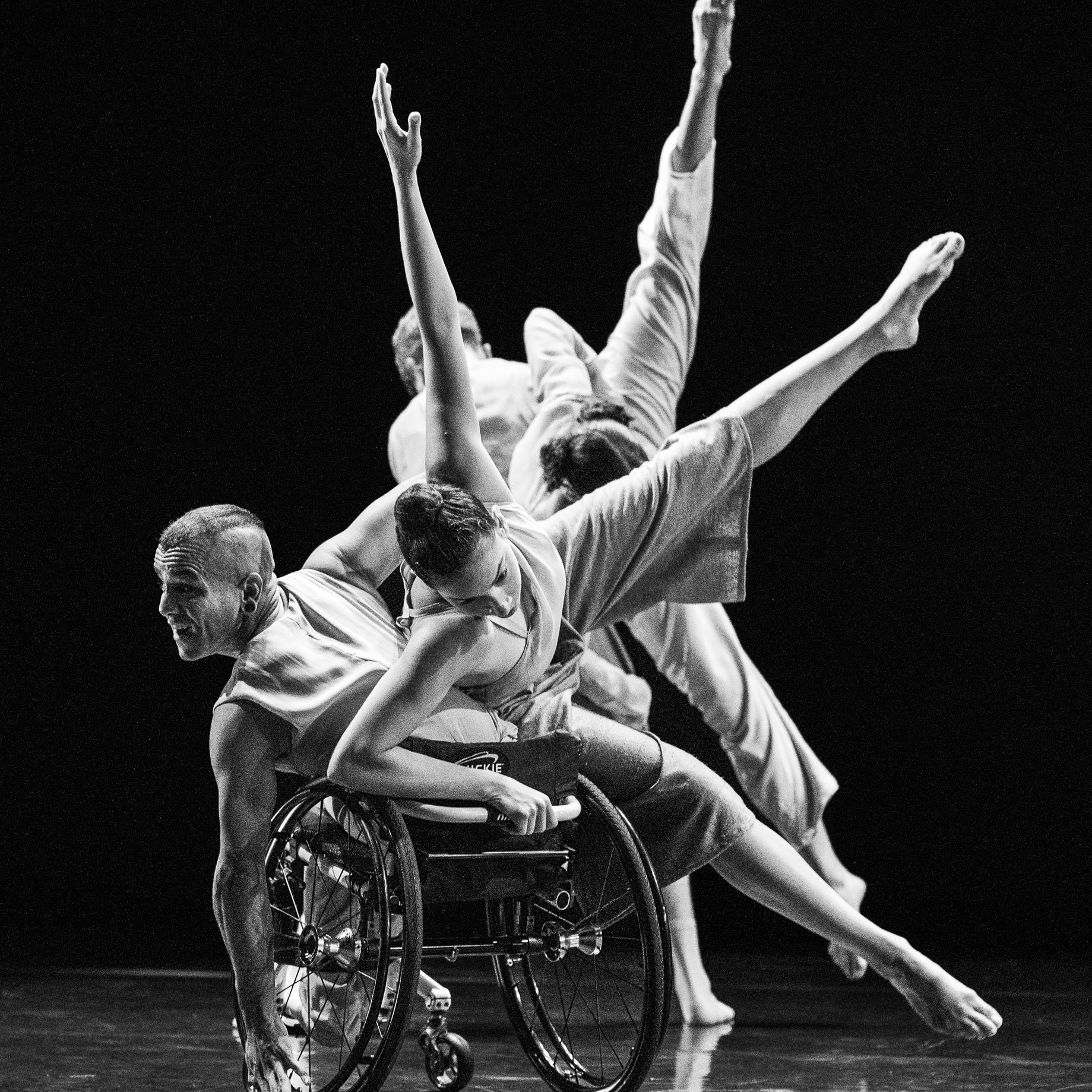In a black and white photo, Janpi tilts their torso to the left in their wheelchair, while Zara balances in a side tilt on Janpi's body & chair with two legs and one arm extended outwards. David and Alaja perform the same movements behind them both.